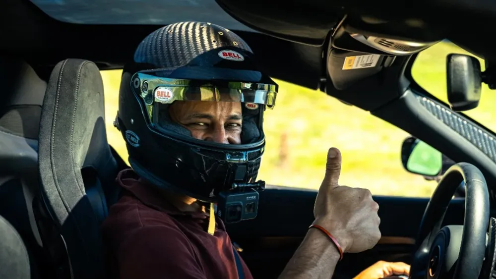 Driver in a race car wearing a Bell helmet with a mounted action camera, giving a thumbs up to the camera/observer.