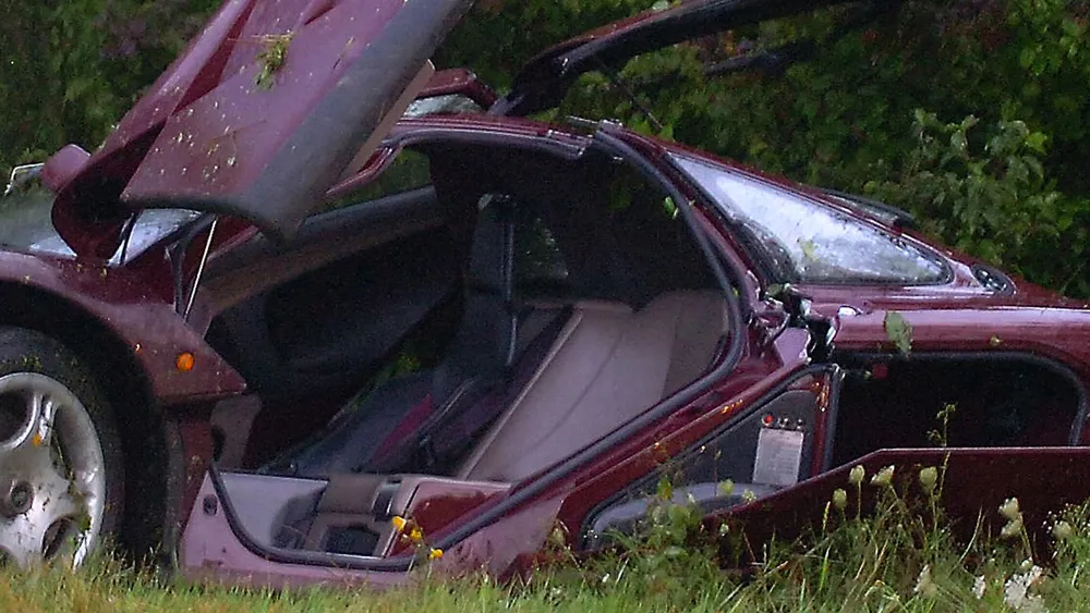 Maroon sports car badly damaged in a grassy area, doors and roof ripped open, interior exposed near a roadside.