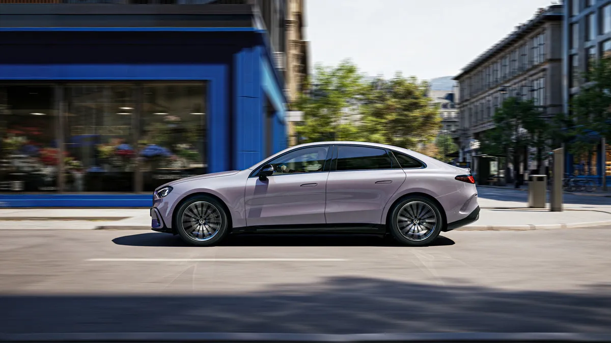 Silver luxury sedan driving down a city street past a blue storefront, with motion blur on the wheels and background side.