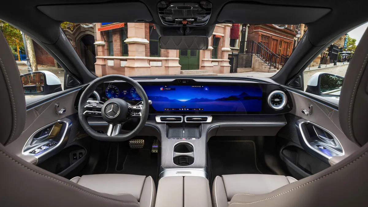 Inside a luxury car: wide curved digital dashboard, steering wheel with Mercedes logo, and tan leather seats.