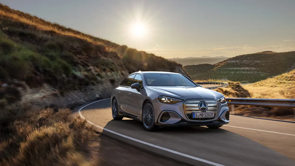 Silver Mercedes-Benz sedan driving on a winding mountain road at sunset with hills and guardrails to the sides.