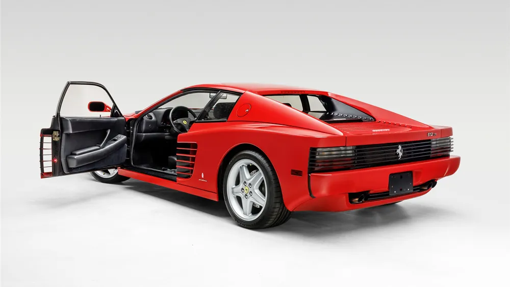 Red Ferrari 512 TR with the driver's door open, black leather interior visible in a white studio setting.