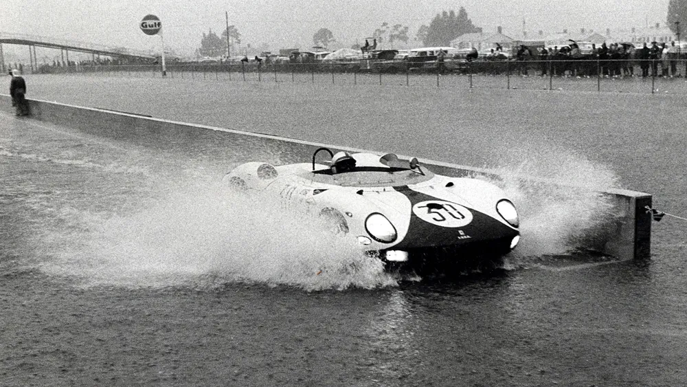 Vintage open-cockpit race car #50 powers through a water barrier, creating a spray as spectators line the track in the background.
