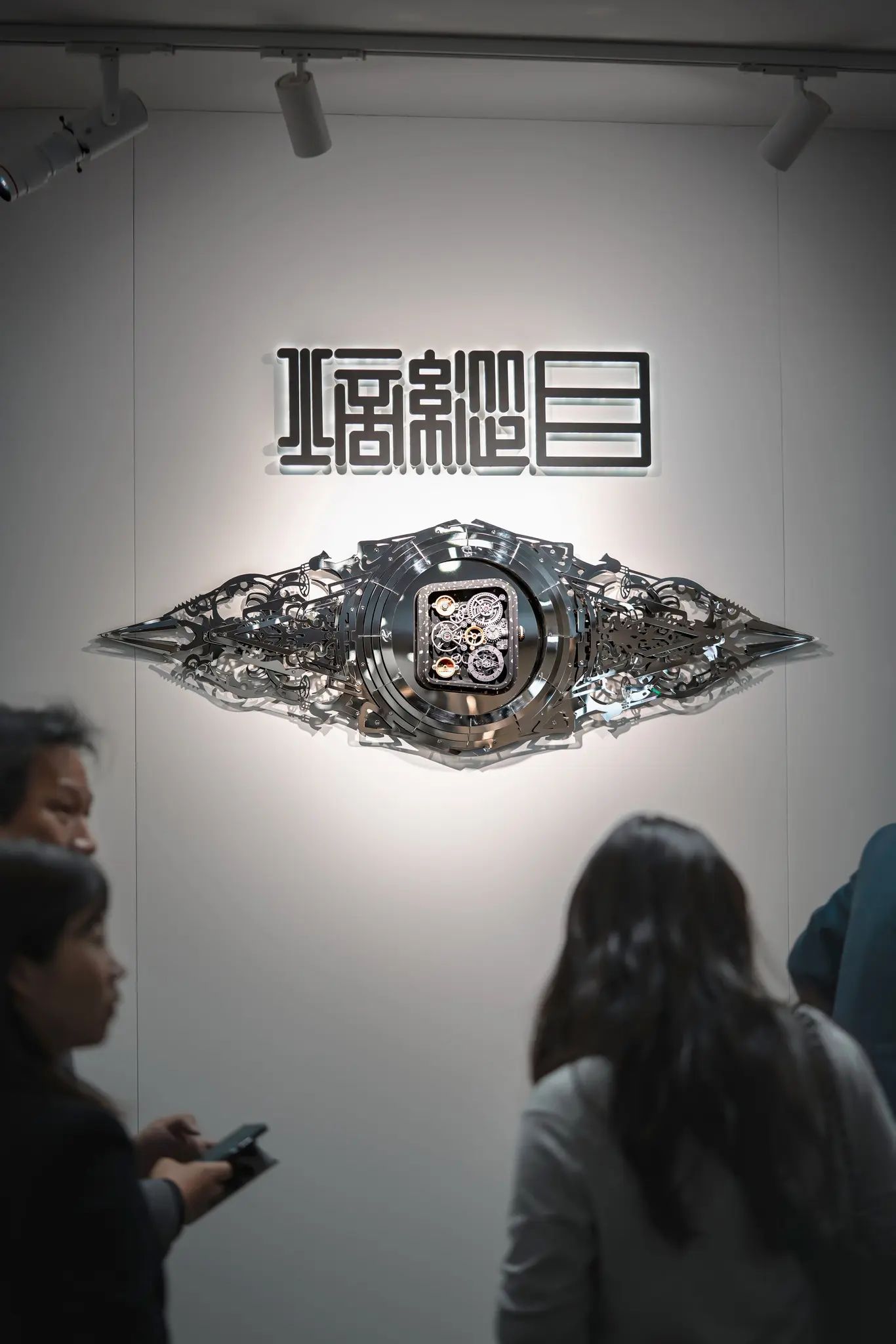 Metallic, gear-filled eye sculpture on a white gallery wall, with illuminated Chinese characters above; a few visitors stand in the foreground.