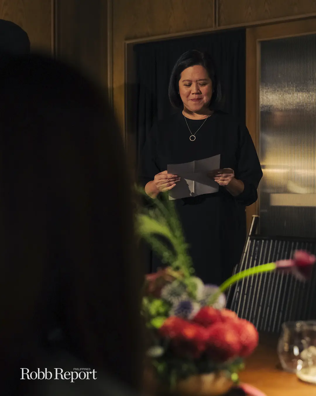 Woman in a black dress reads a folded note at a table with a colorful flower centerpiece during a formal event.