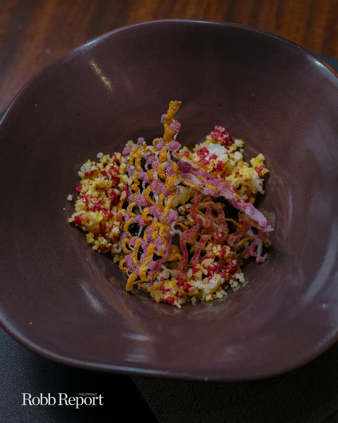Close-up of a plated dish in a dark bowl: a colorful crumb mixture with pink, yellow, and white grains and pinkish-orange crispy strands on top.