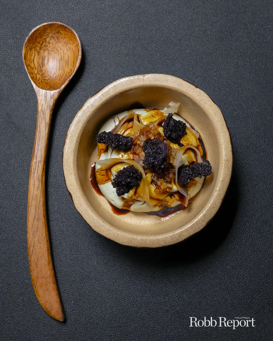 Beige ceramic bowl with a plated gourmet egg dish, topped with onions, dark sauce, and black truffle shards; a wooden spoon rests to the left.