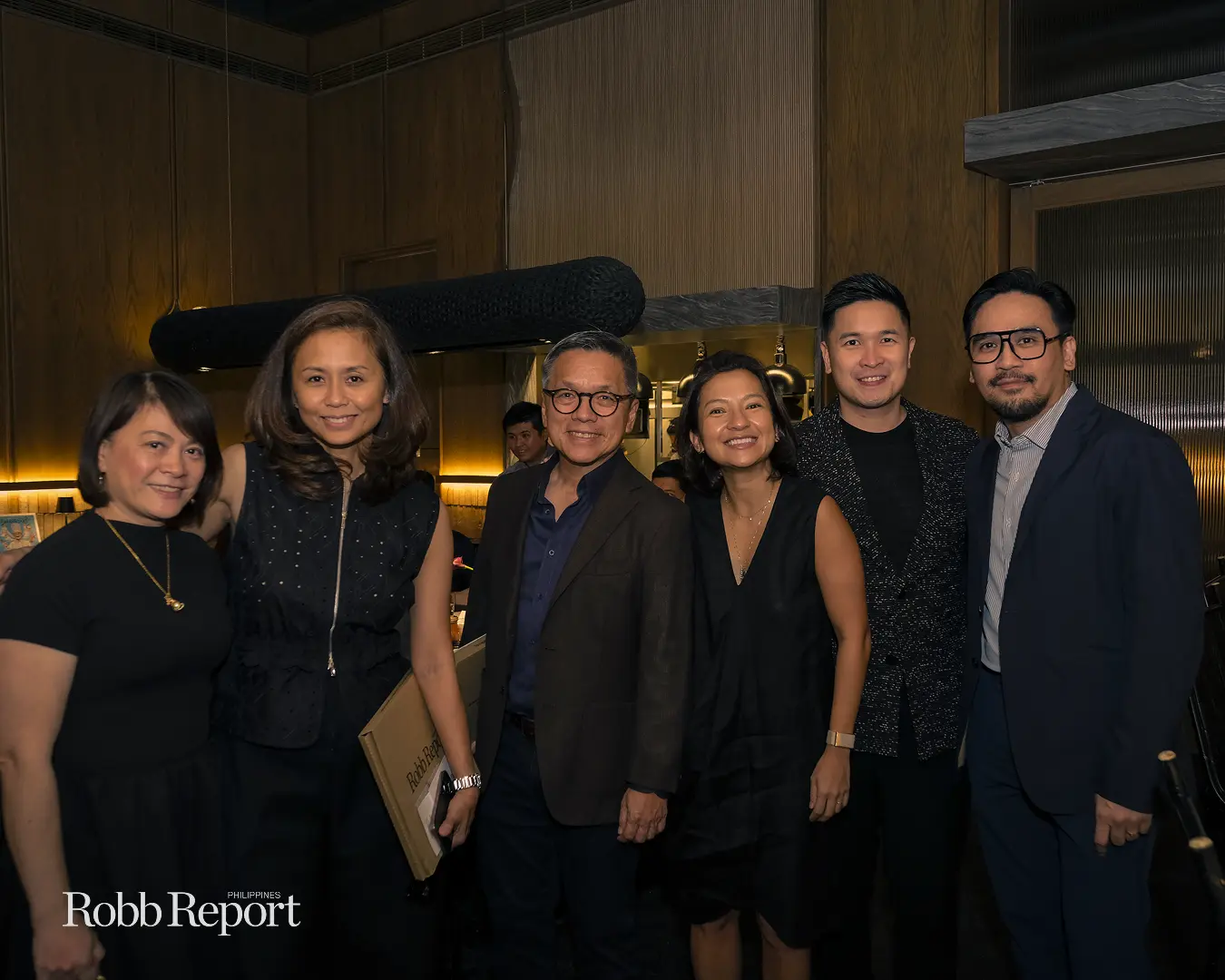 Six well-dressed people posing for a group photo at a dimly lit event, Robb Report Philippines logo visible.