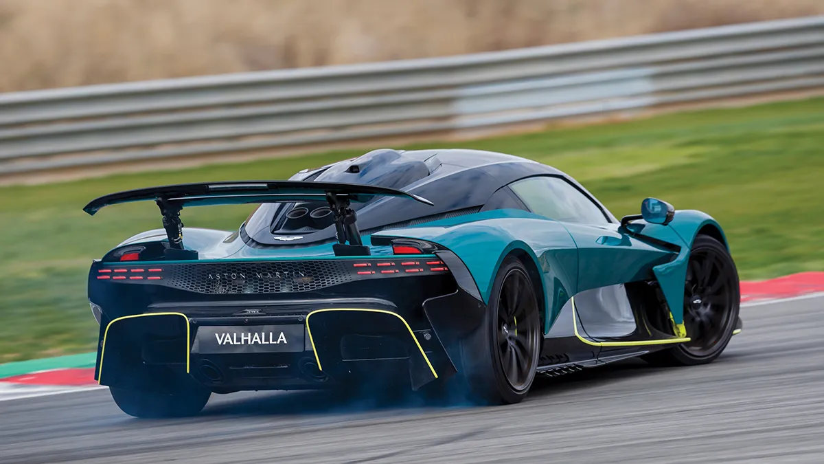 Rear three-quarters view of a blue-teal Aston Martin Valkyrie race car on a track, rear wing up and neon-yellow accents visible.