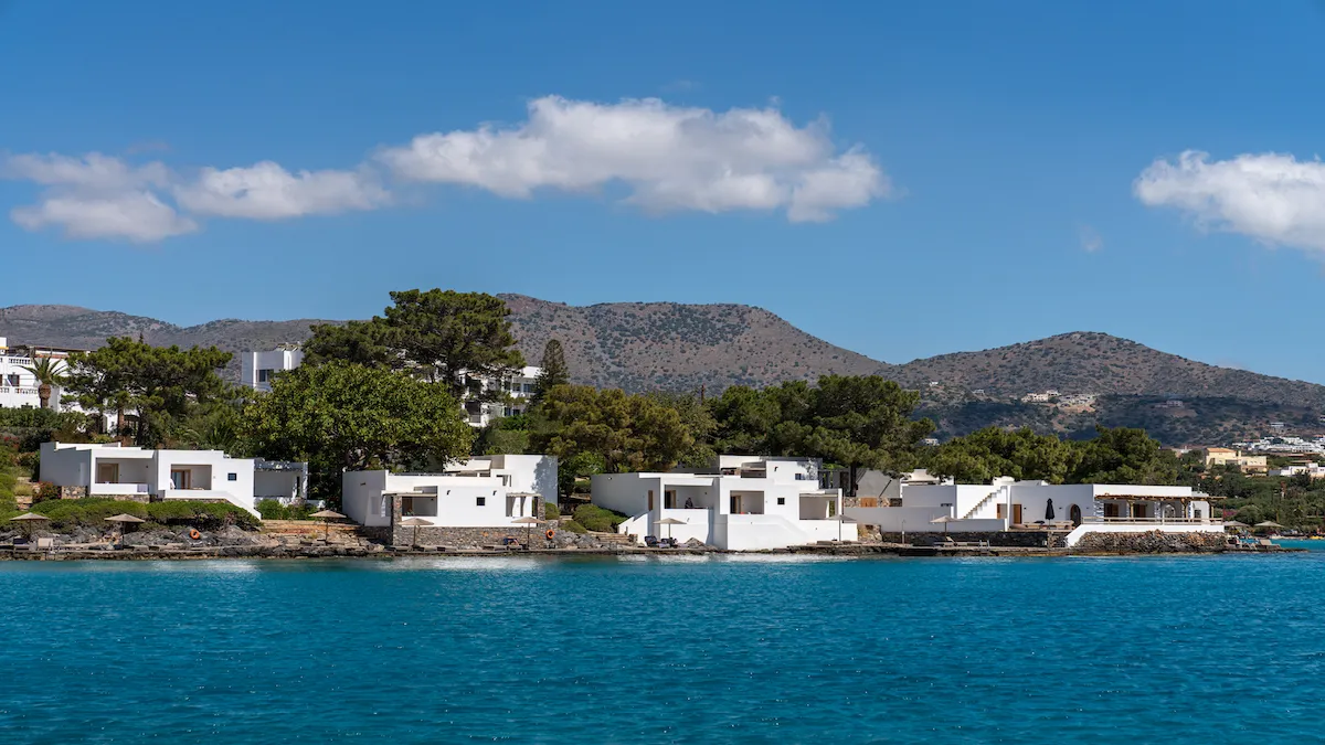 Row of white modern villas along a rocky shoreline, turquoise water in the foreground and hills in the background in a sunny coastal scene.