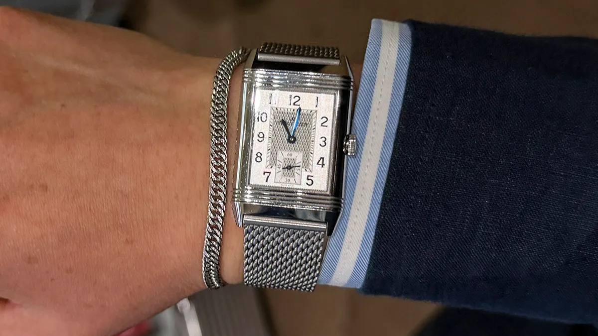 Silver rectangular wristwatch with a mesh band on a man's wrist, beside a silver bracelet, under a blue suit cuff.