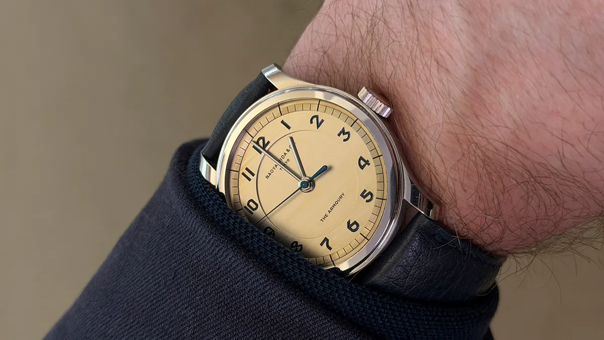 Close-up of a vintage-style analog wristwatch with a beige dial, blue hands, black numerals, and a black leather strap on a wrist wearing a dark sleeve.