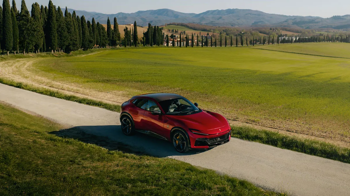 Red sport SUV cruising along a narrow rural road with green fields, tall cypress trees, and distant hills.