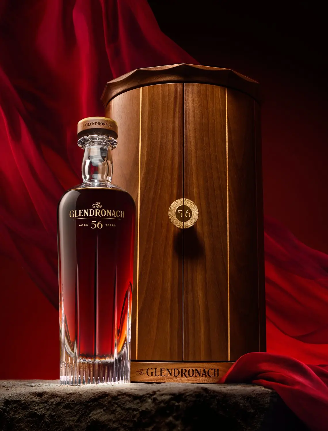 The Glendronach 56-year whiskey bottle beside a wooden display cask against a red velvet backdrop.