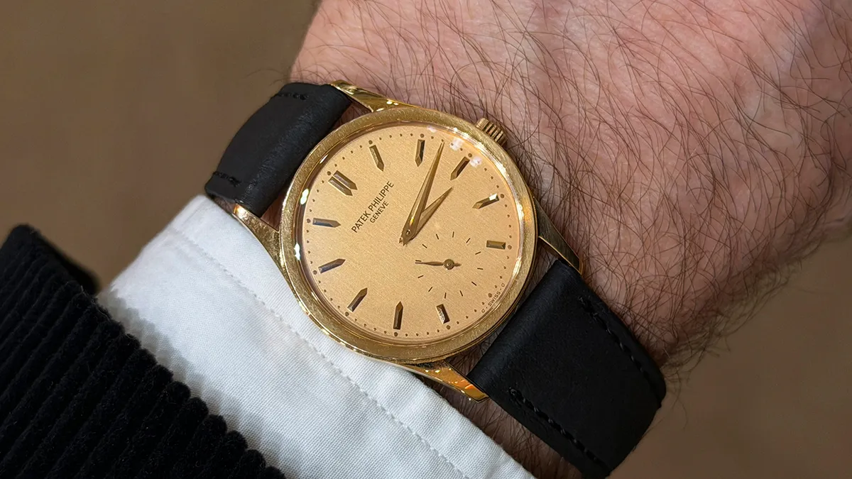 Gold Patek Philippe wristwatch on a black leather strap, worn on a light-skinned wrist with cuffed shirt sleeve visible.