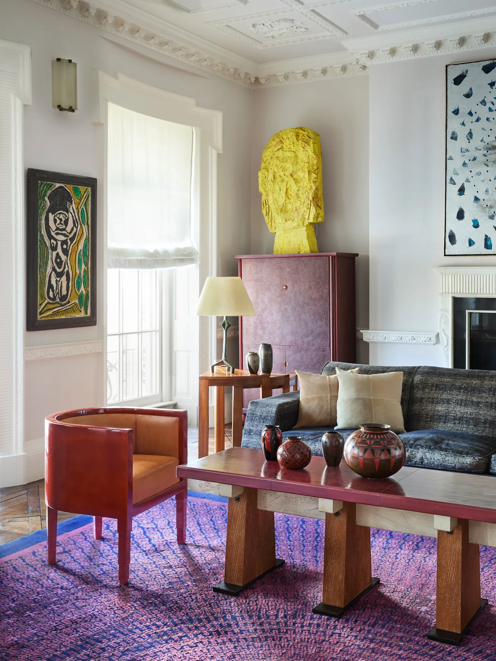 Bright living room with a bold yellow sculpted head on a pink cabinet, next to a gray-blue sofa and a wooden coffee table displaying red and black vases, atop a purple patterned rug.