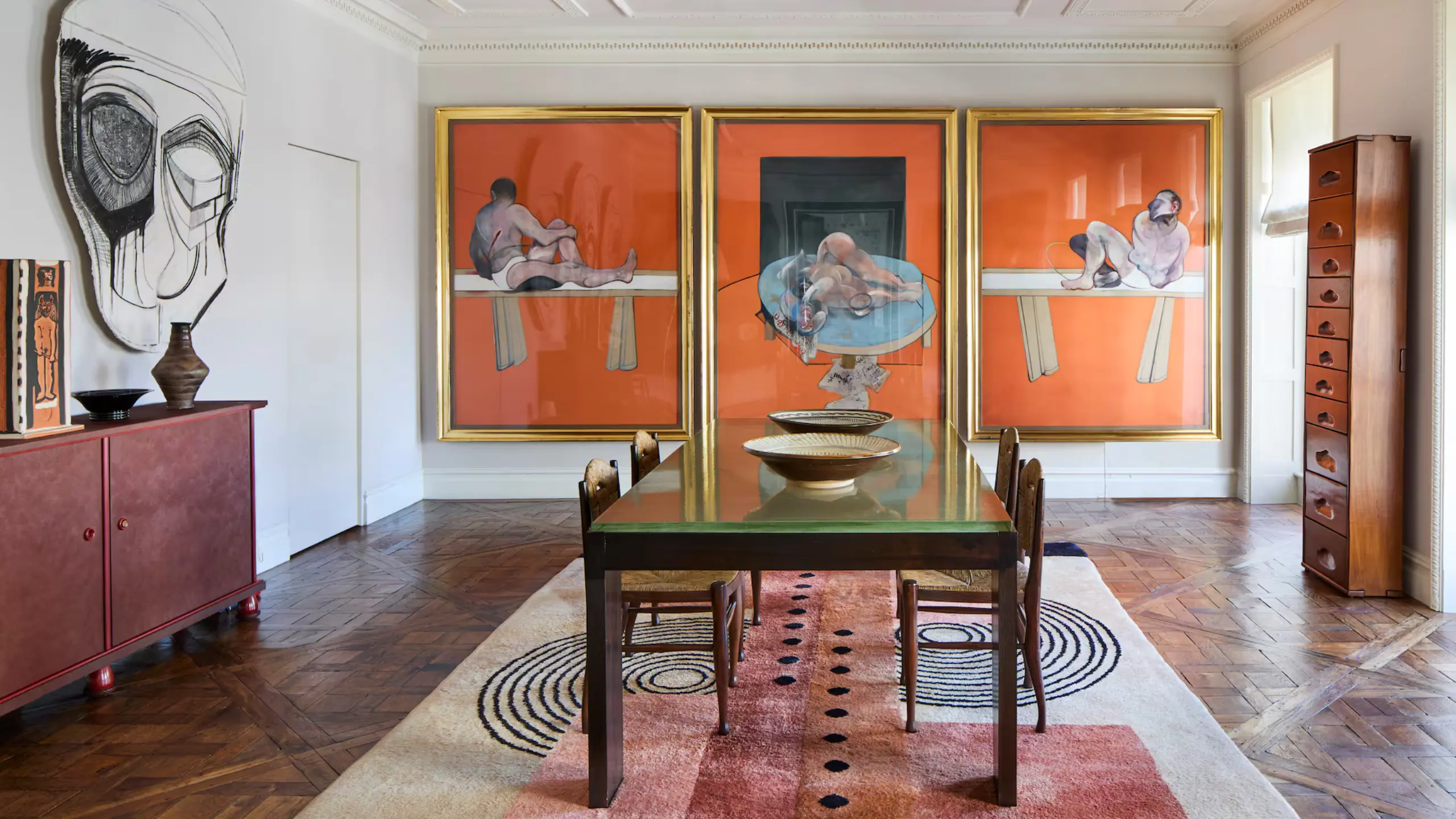 Dining room with a glass-topped table, wooden chairs, and three orange-framed paintings along the back wall.