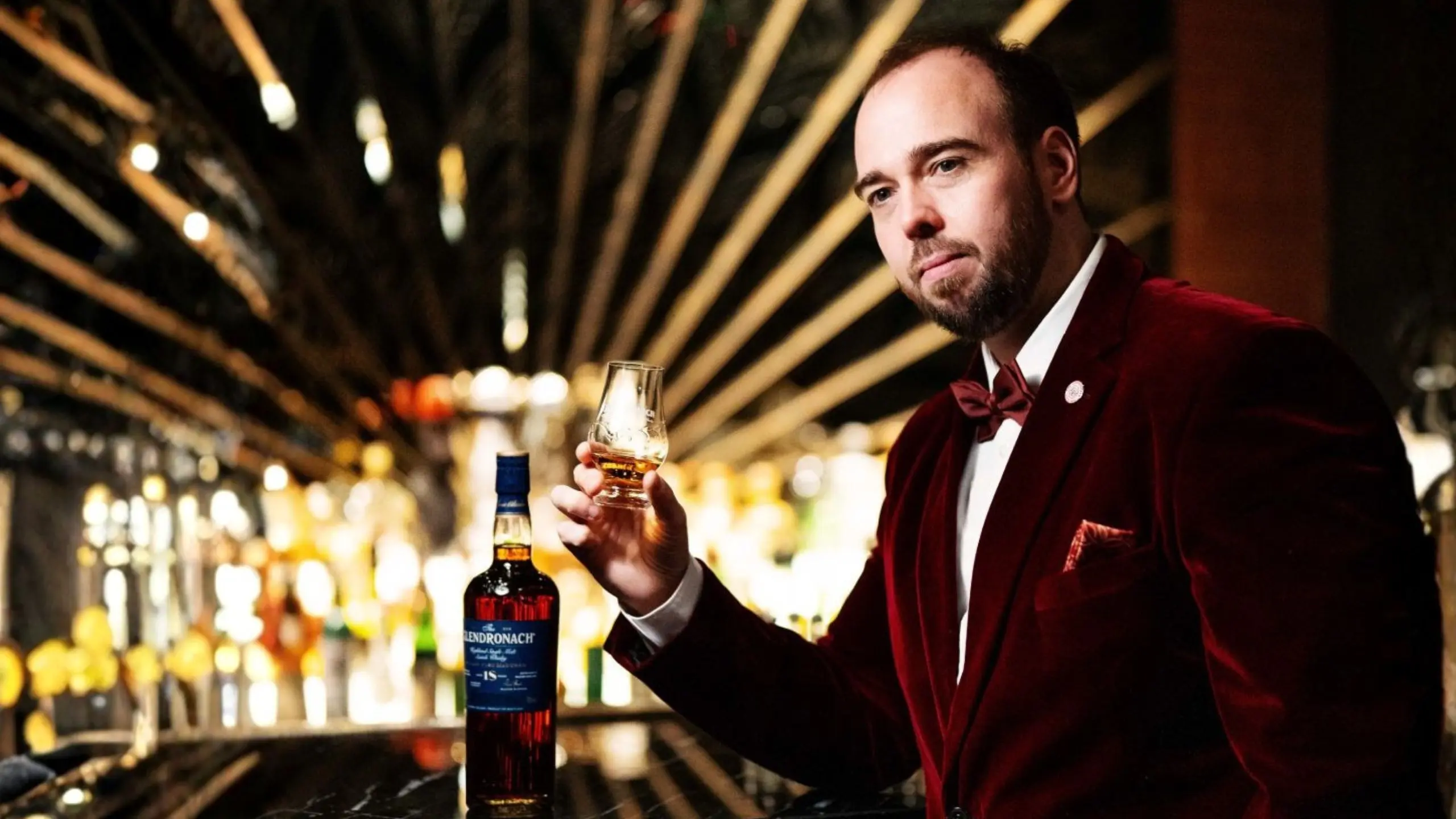 Bartender in a burgundy velvet suit pours whiskey while holding a glass at a dim bar with backlit bottles behind him