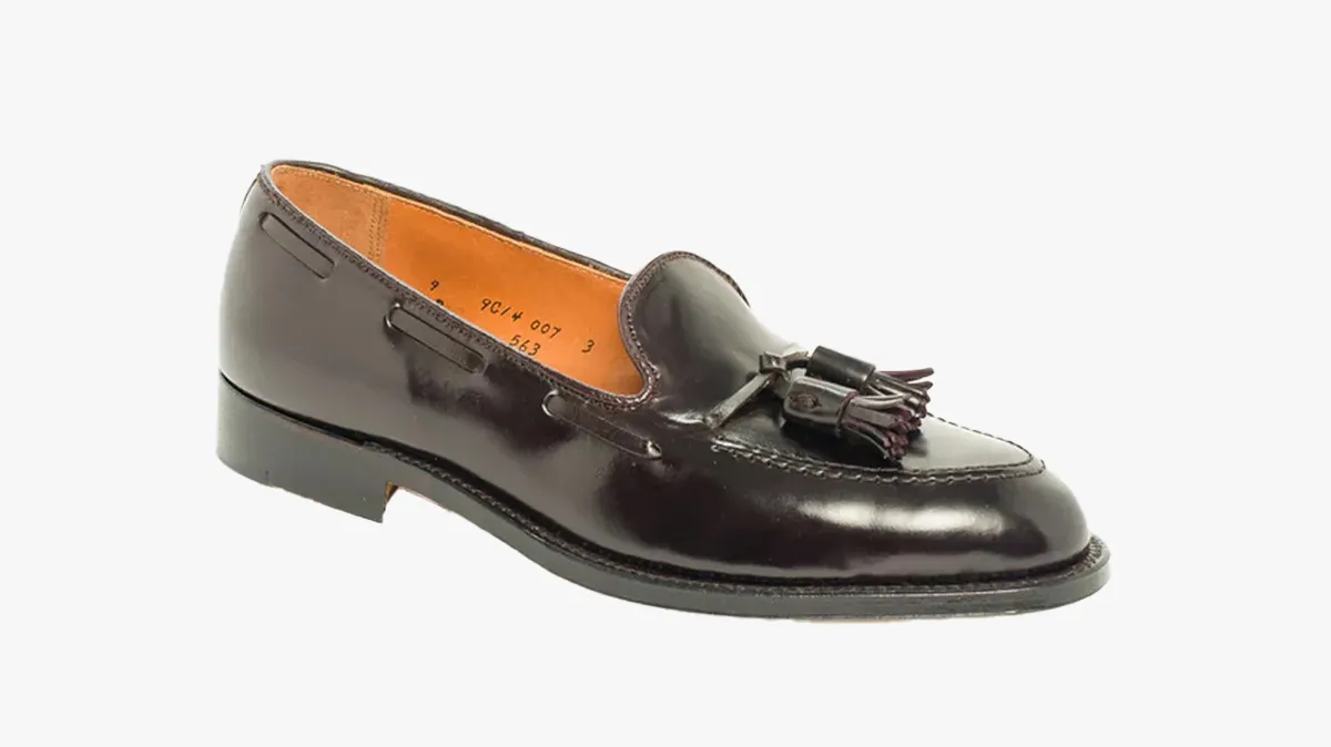 Black patent leather tassel loafer with a low black sole, shown in profile against a white background, interior tan lining visible.