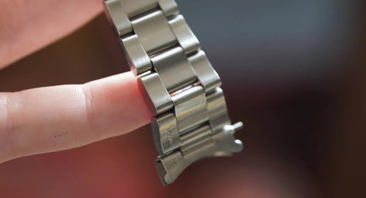 Close-up of a metal watch bracelet being held by a finger, with a pin emerging between links into view.