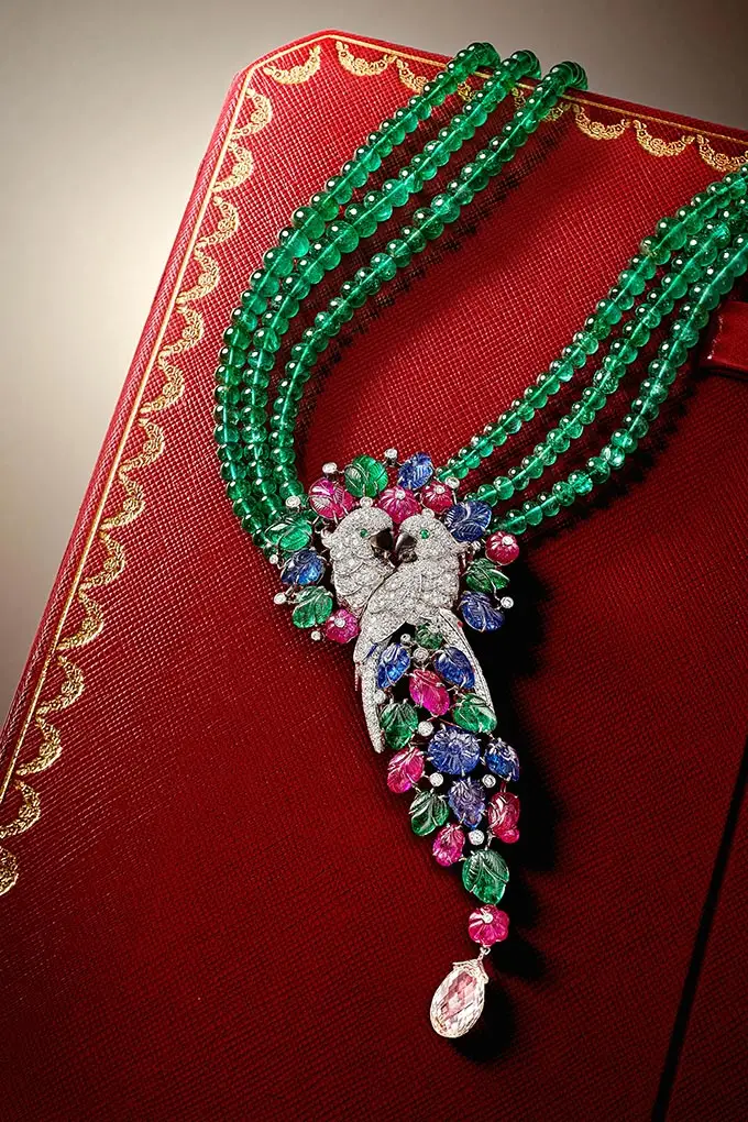 Green beaded multi-strand necklace with a diamond-encrusted bird pendant on a red textured box; colorful gem accents and a teardrop pendant.