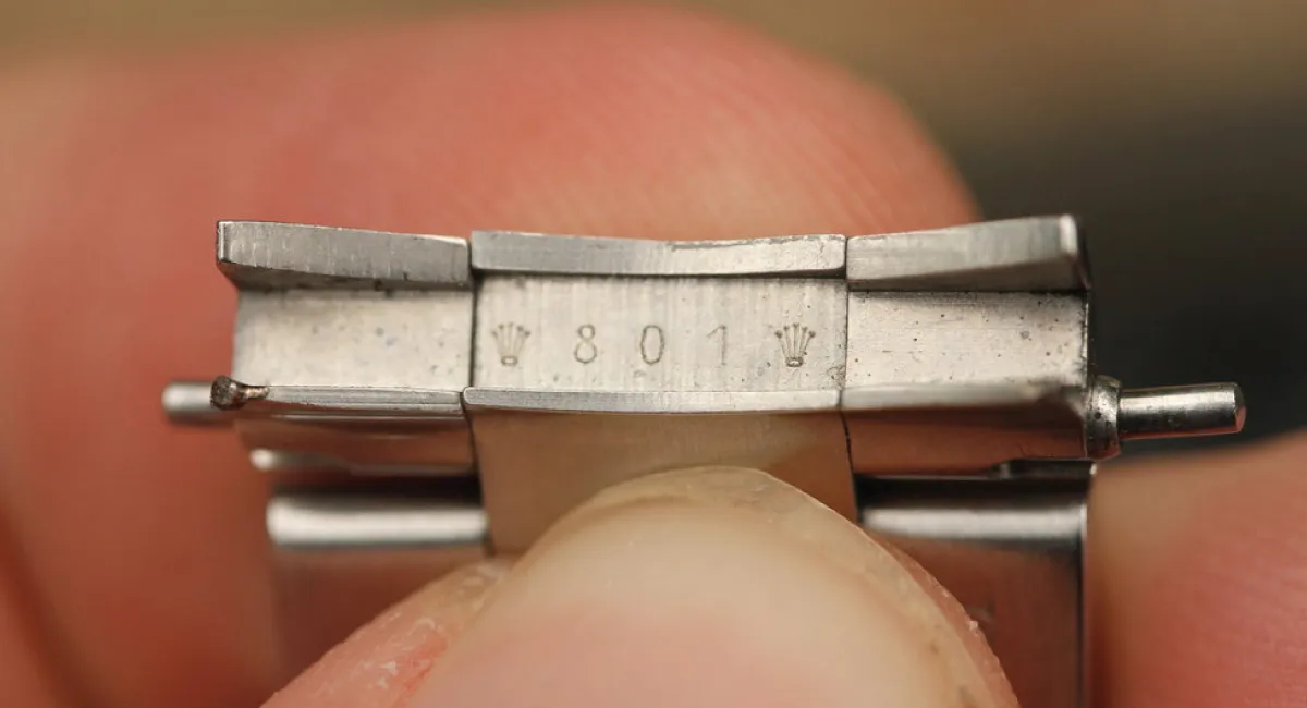 Close-up of a metal watch clasp held by fingers, showing engraved '801' and small crown marks.