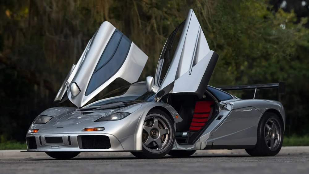 Silver exotic sports car with butterfly doors raised, revealing red interior, parked outdoors on a paved surface.