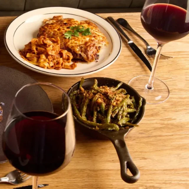Tabletop dinner spread: beef steak on a gray plate, baked pasta, sautéed green beans in a skillet, a corn dish, and two glasses of red wine on a wooden table.