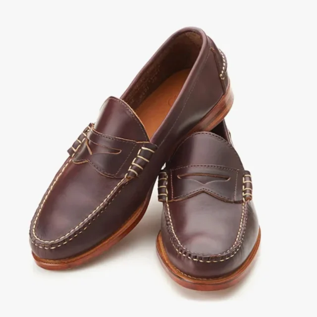 Four men's loafers floating on a green background: black penny loafer with gold horsebit, black tassel loafer, black cap-toe loafer, and dark brown suede loafer.