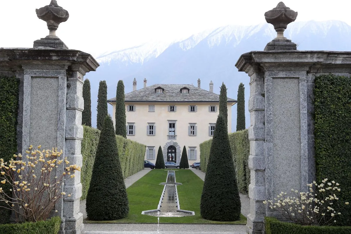 Stately villa framed by stone pillars and topiary, with a central fountain leading to the entrance amid a manicured garden and distant mountains.