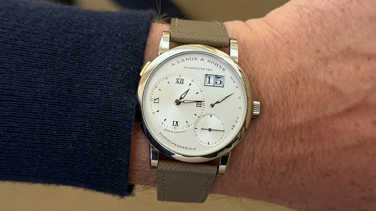 Silver A. Lange & Söhne wristwatch with white dial, Roman numerals, large date window at 1–2 o'clock, and small seconds subdial on a brown leather strap, worn on a wrist.