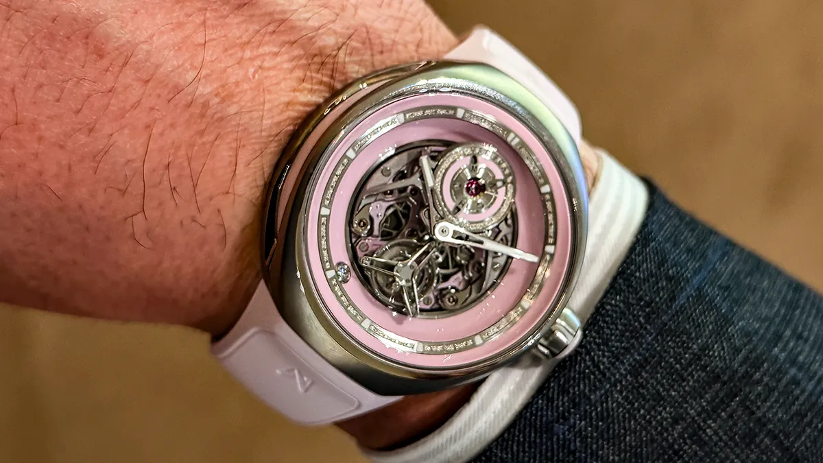 Skeleton wristwatch with pink dial and exposed gears on a white strap, worn on the wrist.