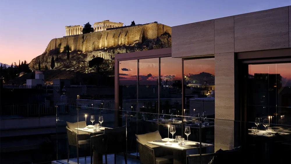 Rooftop restaurant at dusk with glass railing, overlooking Athens; the illuminated Acropolis sits on a hill in the distance.