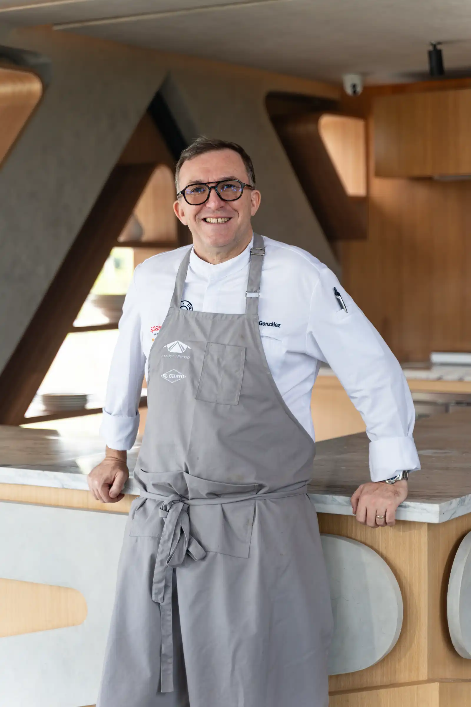 Chef Chele Gonzalez in a white uniform and gray apron leans on a marble counter in a modern wooden kitchen, smiling at the camera.
