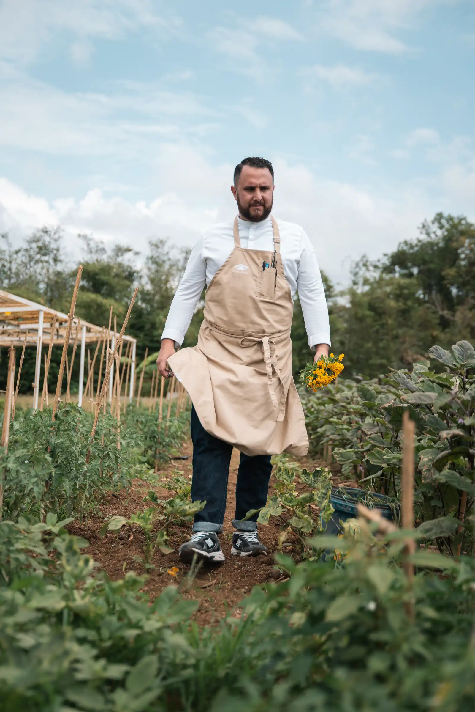 Chef Rodrigo Osorio in a beige apron holding a bouquet of yellow flowers in a vegetable garden on a sunny day.