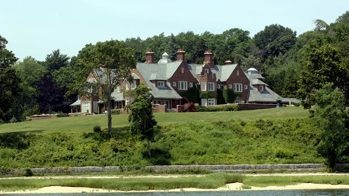 Brick manor with multiple chimneys and gabled slate roofs on a manicured hilltop, surrounded by trees by the water.