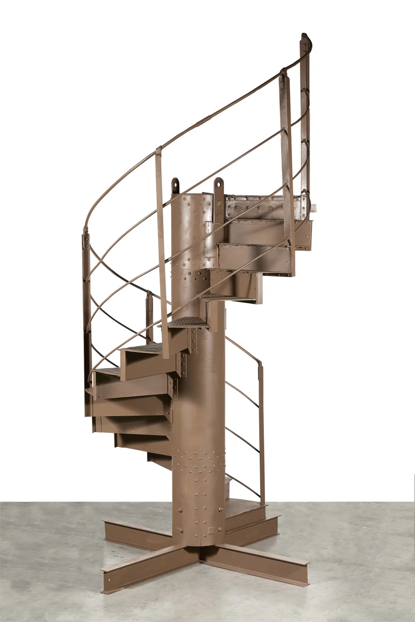 Brown industrial spiral staircase with riveted steel posts, curved railing, and a central column on a concrete floor.