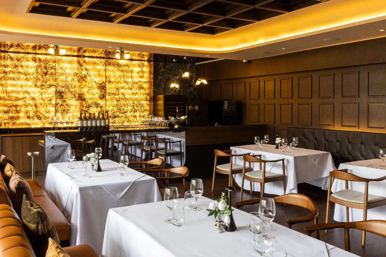 Elegant dining room with white-clothed tables, wine glasses, and wooden chairs in warm ambient lighting.
