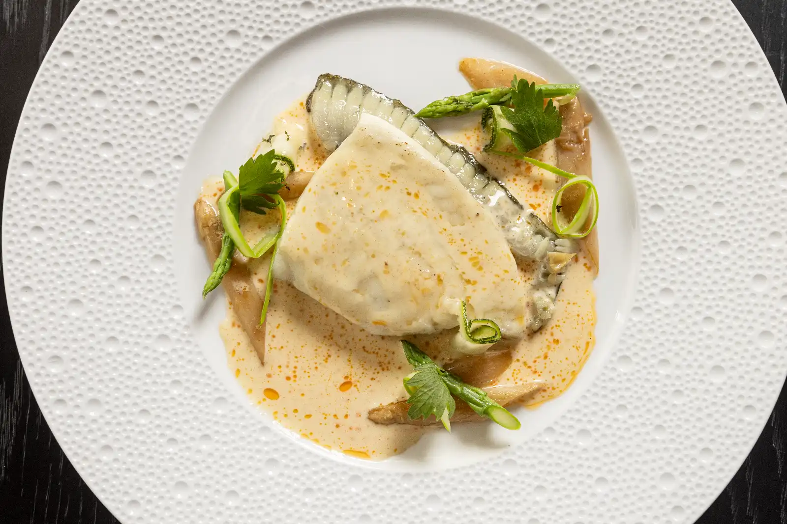 Plated white fish fillet in creamy orange sauce, garnished with greens and herbs on a textured white plate