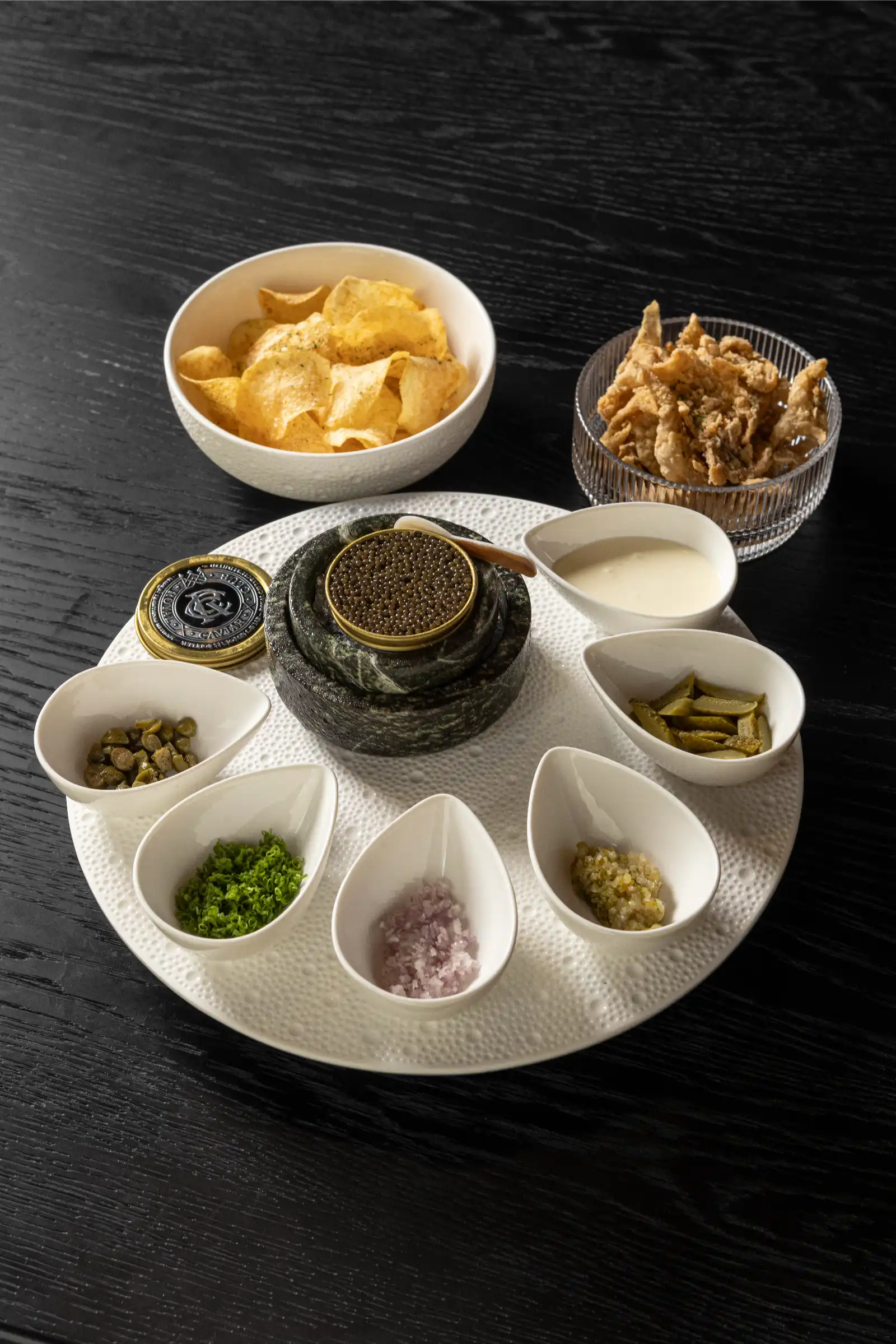 A round white serving platter with a black tin of caviar in the center, surrounded by small bowls of chopped herbs, pickles, onions, sauces, chips, and a side dish of fried items.