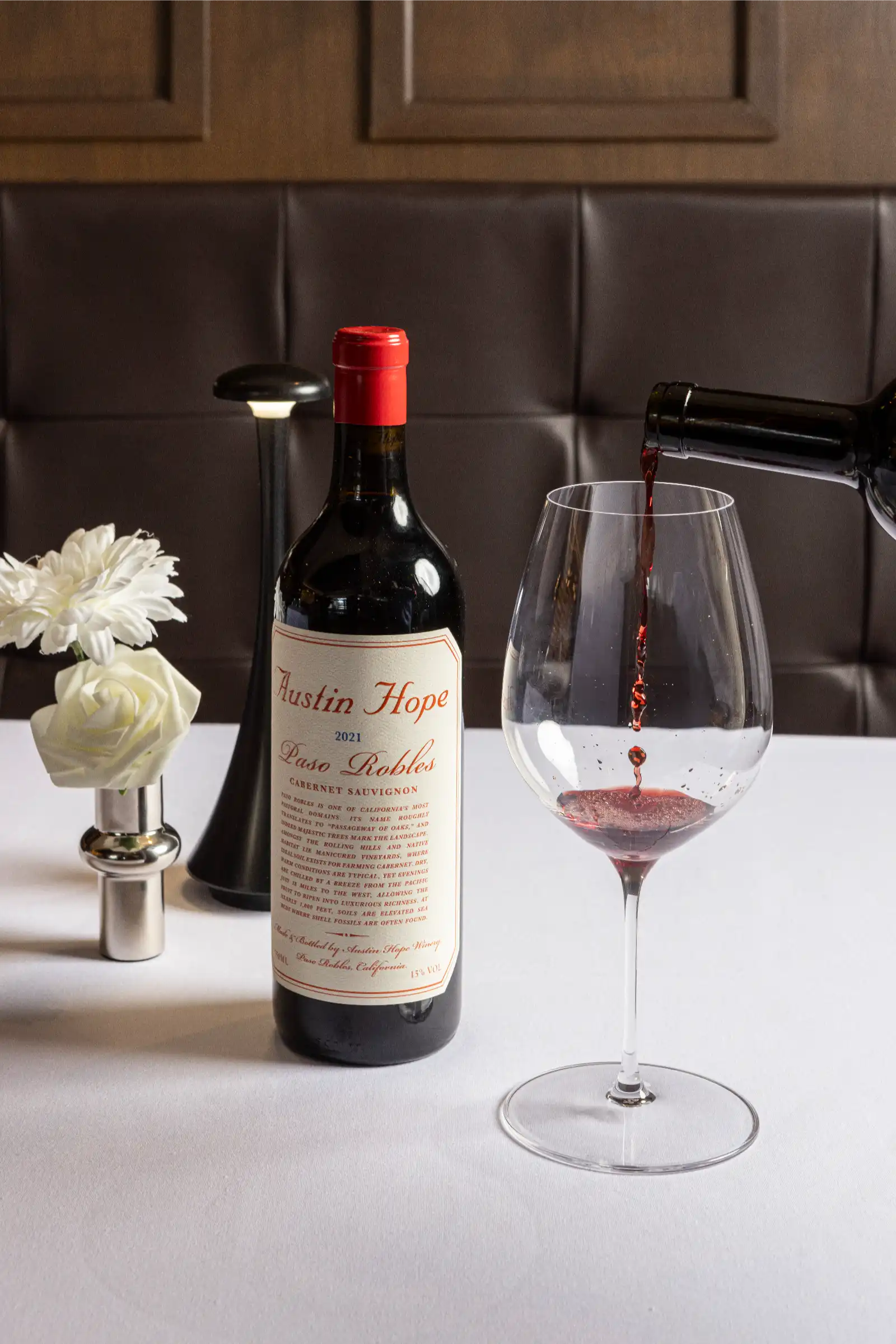 Bottle of Austin Hope 2021 Cabernet Sauvignon on a white table, red wine being poured into a stemmed glass, with a white flower in a silver vase to the left and a dark, padded backdrop behind.