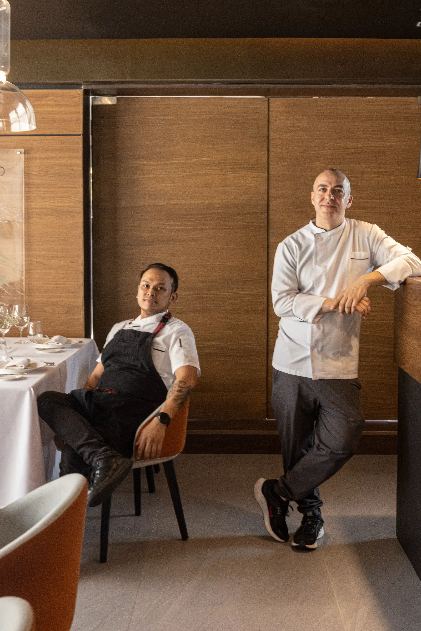 Two chefs pose in a dining area: one seated in a black apron, the other standing with arms crossed against a wood-paneled wall.