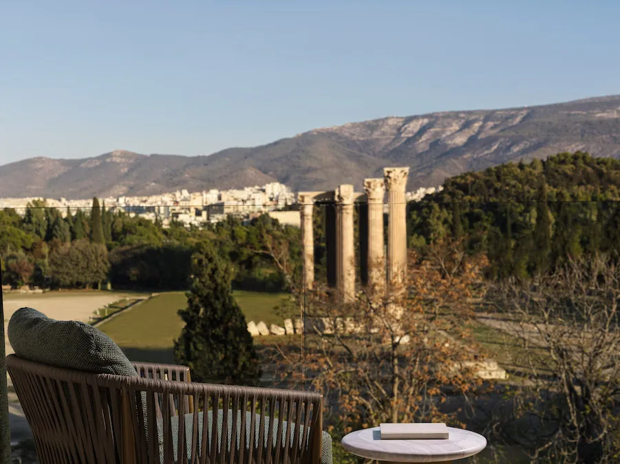 Outdoor patio chair with cushion on a deck, facing ancient stone columns amid trees and distant mountains.