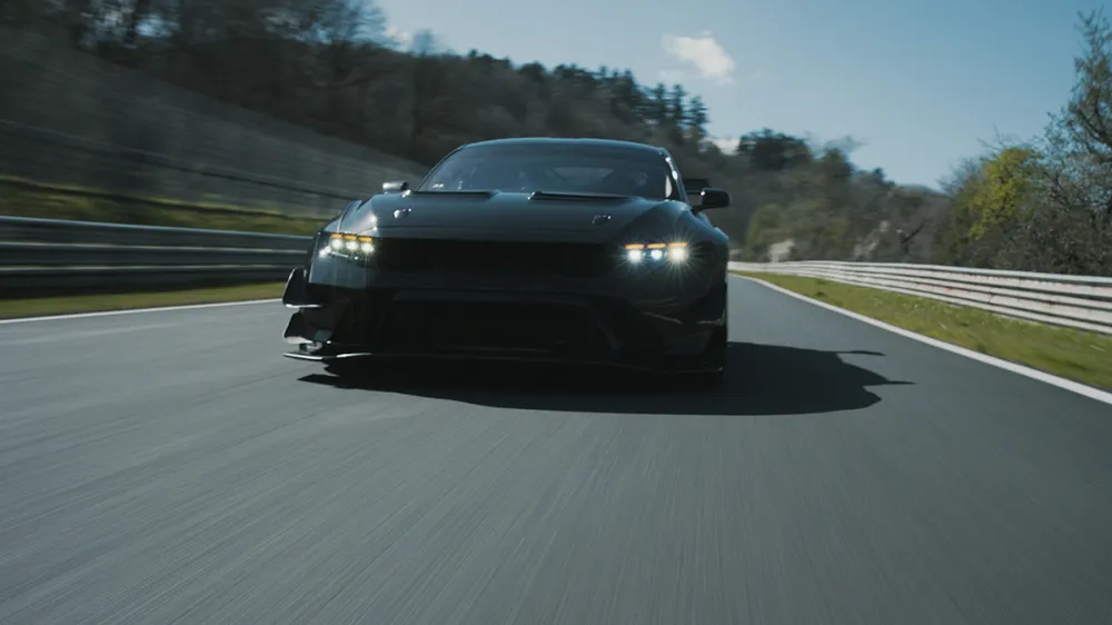 Dark sports car speeds toward the viewer on a race track, headlights on, with blurred surroundings.