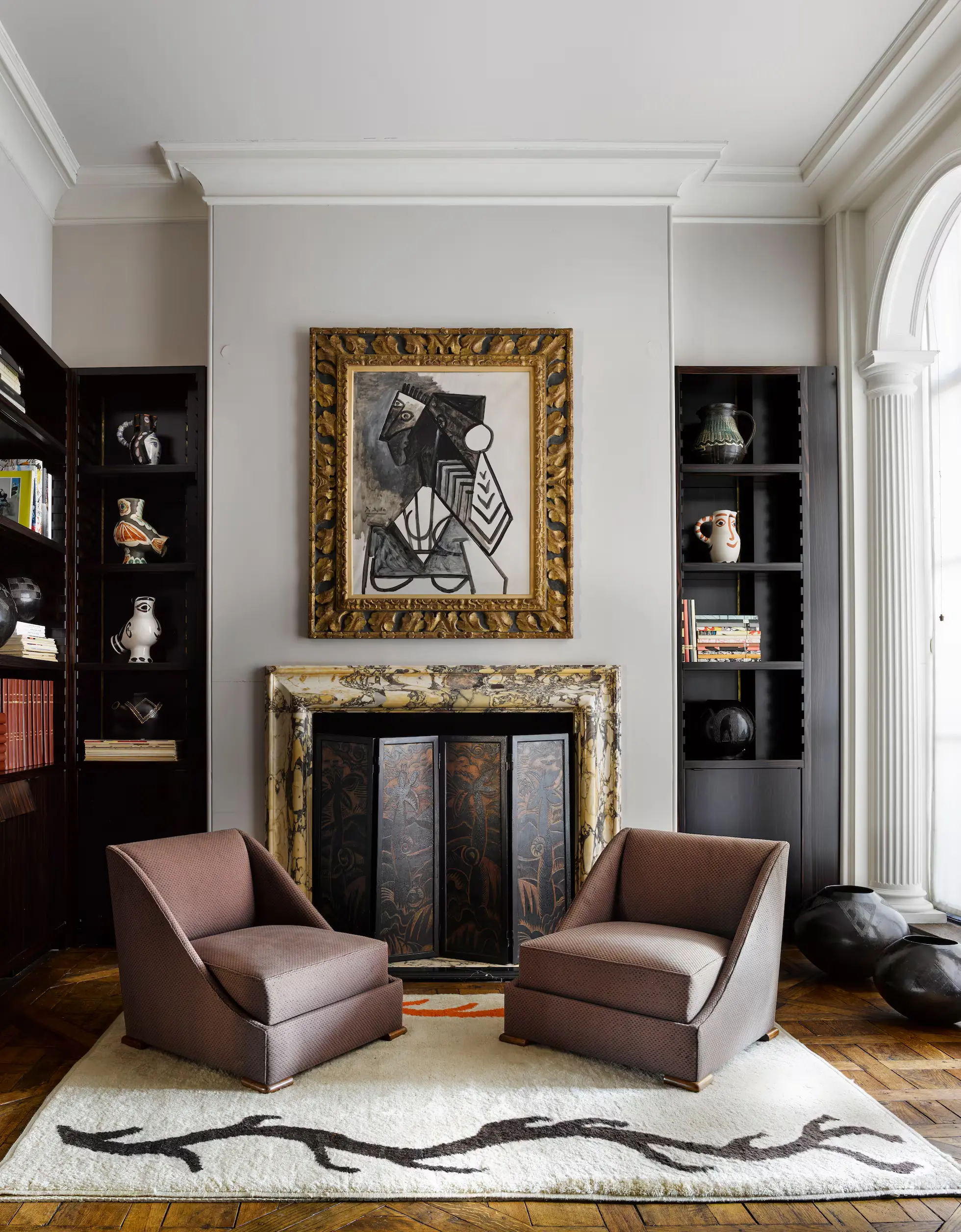 Living room seating area with two mauve armchairs facing a gilded fireplace and an abstract framed artwork above it in a tall, stylish room.