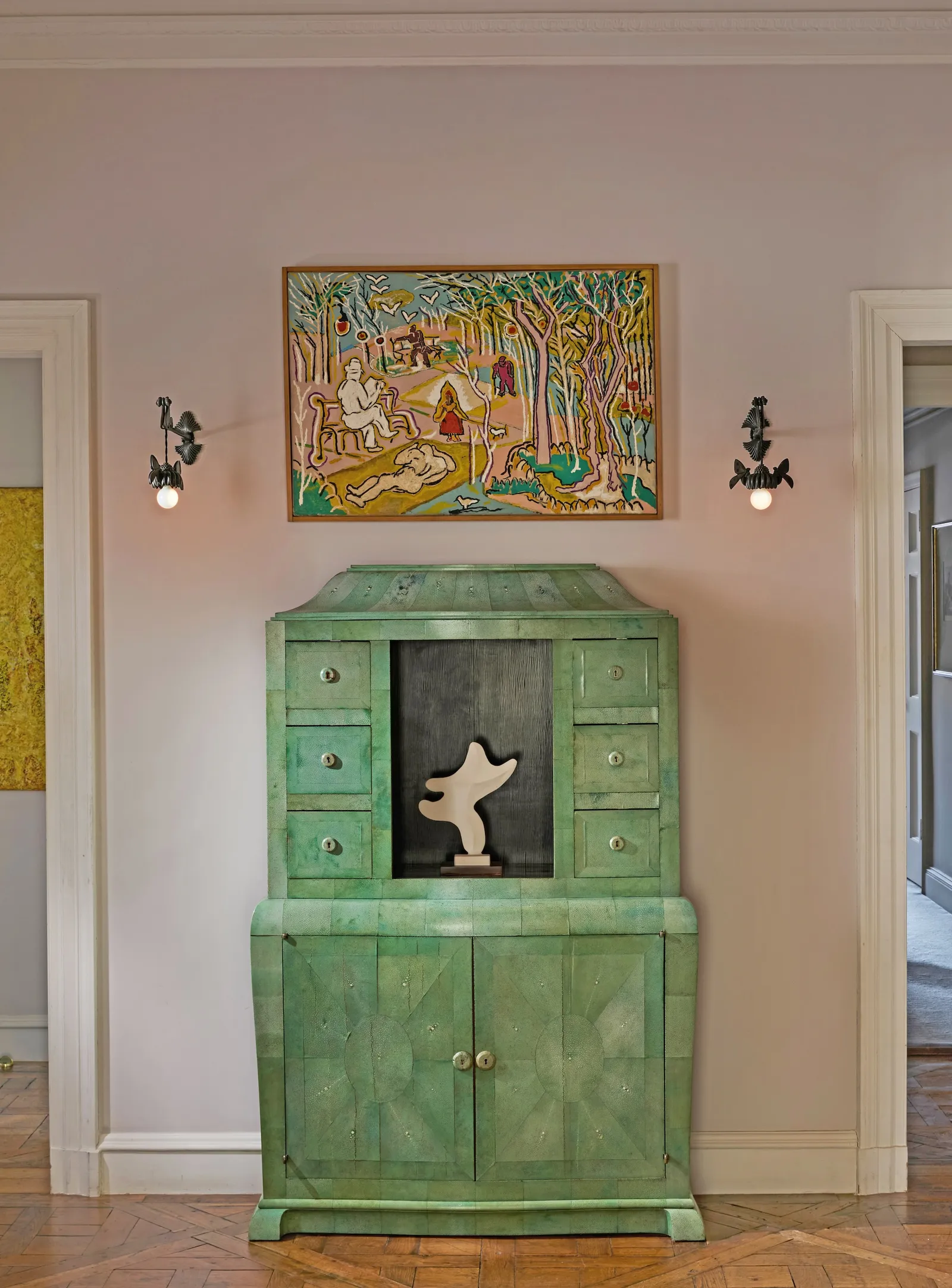 Green antiqued cabinet with drawers and panel doors sits against a pale pink wall, a white abstract sculpture in the center niche, and a colorful painting above.