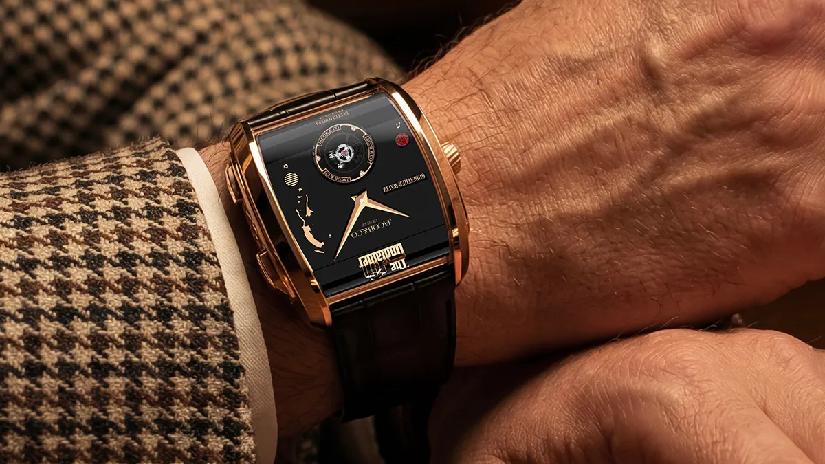 Rose gold rectangular wristwatch on a man’s wrist, black dial with gold hands, worn over a houndstooth-patterned jacket cuff beside a white shirt cuff.