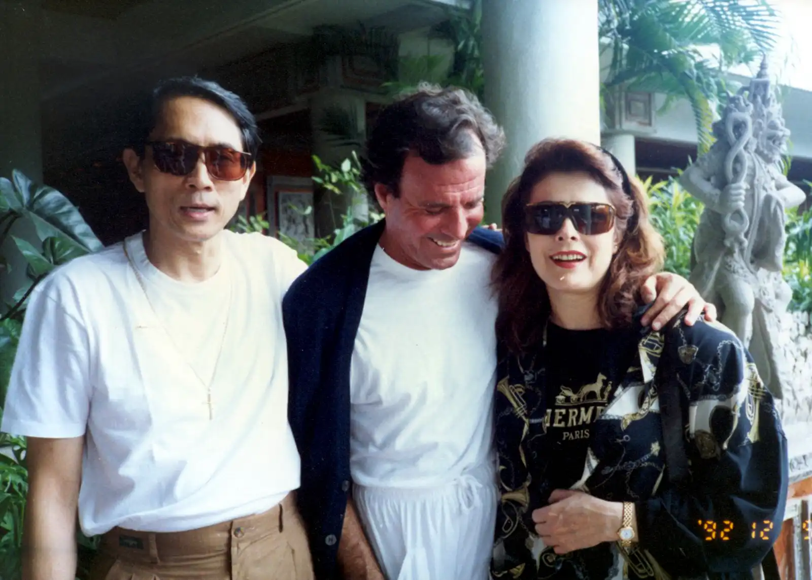 Three adults posing together outdoors, wearing sunglasses; left man in white T‑shirt and cross necklace, center man in white outfit with dark jacket, right woman in a patterned jacket.