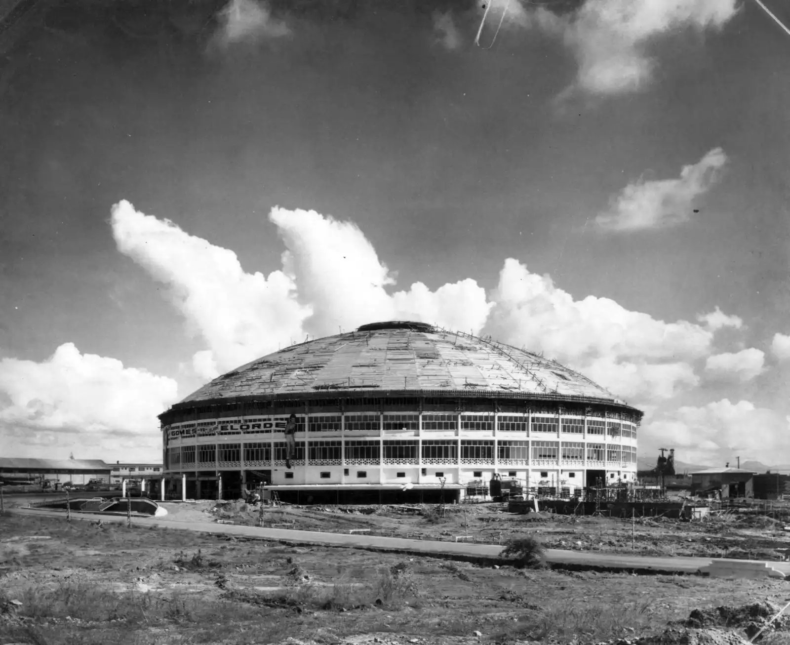 Under-construction circular domed building with scaffolding around the exterior against a cloudy sky.