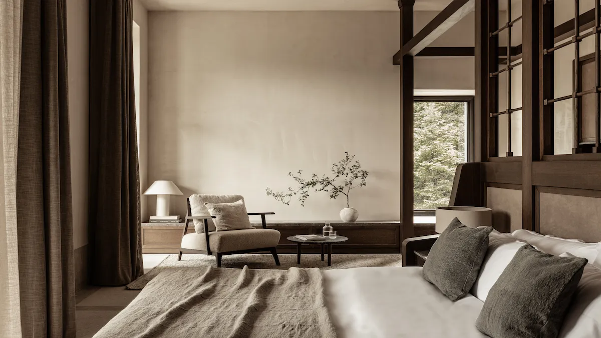Neutral-toned bedroom with a seating area: cream armchair, small round coffee table, and a low wooden cabinet by a large window.